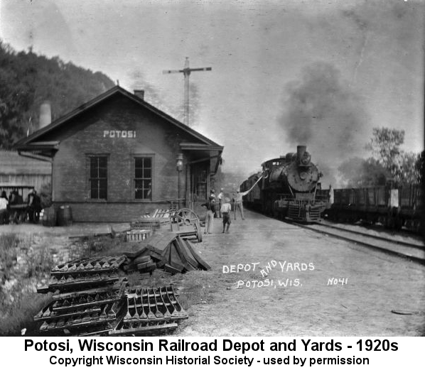 Black and white photo of a small railroad depot building with a steam locomotive puffing smoke on the nearby tracks; workmen stand between the depot and the locomotive. Behind them stand a steep wooded hill and an antenna tower under a grey sky.